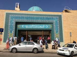 The outside of Central Mosque in Lisbon, a sandy brick building with striking aqua tile work around the entrance.g with There are people gathered and moving about in front, two cars parked on the street.