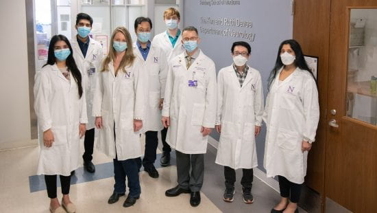 Koralnik lab members stand grouped together wearing white lab coats and PPE