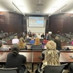 A view of students from the back looking out to a group of panelists speaking in a lecture hall