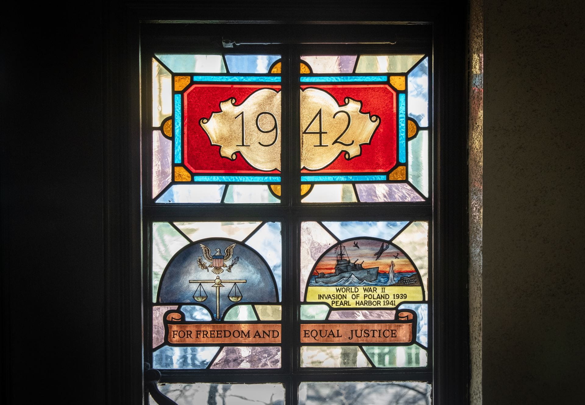 The 1942 stained glass includes the scales of justice side by side with a battleship and plane against a sunset or sunrise, and the text 