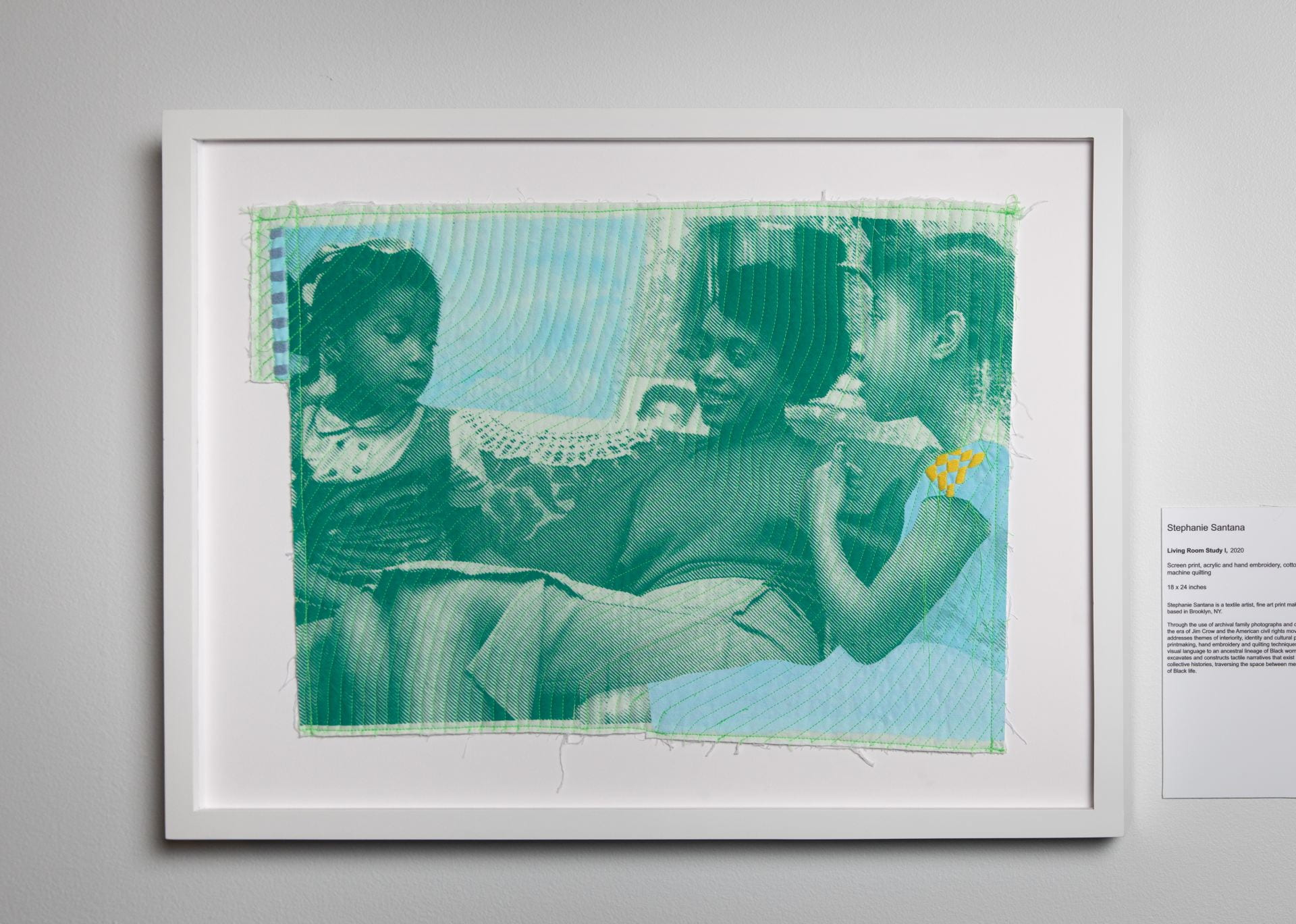 A black woman sitting between two black children, all printed on green and blue quilted cloth with yellow embroidery on one child's dress. There is a doiley on the back of the floral couch that the girls are sitting on. 