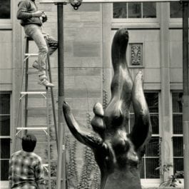 Man on a ladder installing Richard Hunt's statue, Why? in the Law School's courtyard. The statue is comprised of 6 long, undulating forms reaching upwards from a square base.