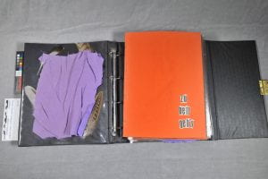 a black 3-ring binder with a purple piece of cloth on the left side and an orange piece of paper on the right.