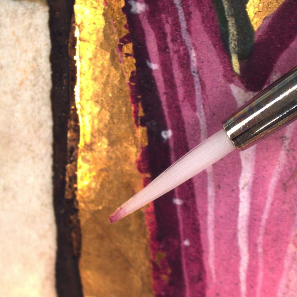 A small brush with red pigment on the end hovering over gold and magenta paint.