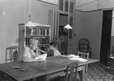 One soldiers reads at a table while another looks for a volume in the bookcase