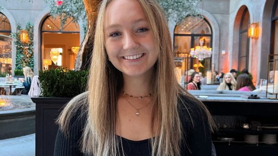 Georgia sits smiling at a restaurant wearing a black shirt and gold necklaces